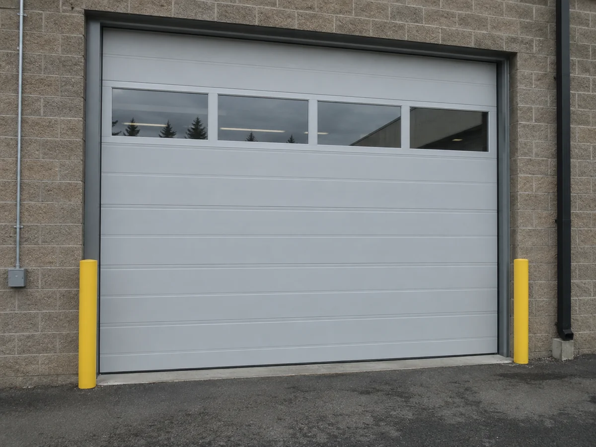 Light-gray industrial sectional door with upper windows and yellow protective posts.